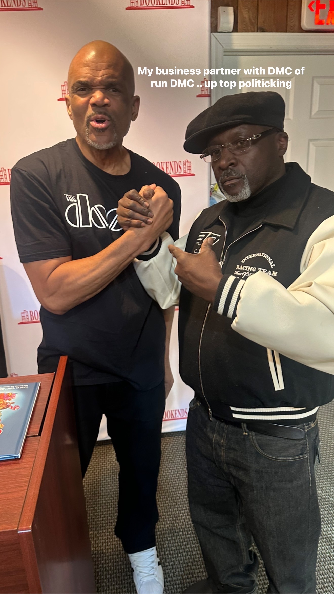 Two Men in Casual Clothing Pose Together Indoors Clasping Hands and Looking at the Camera