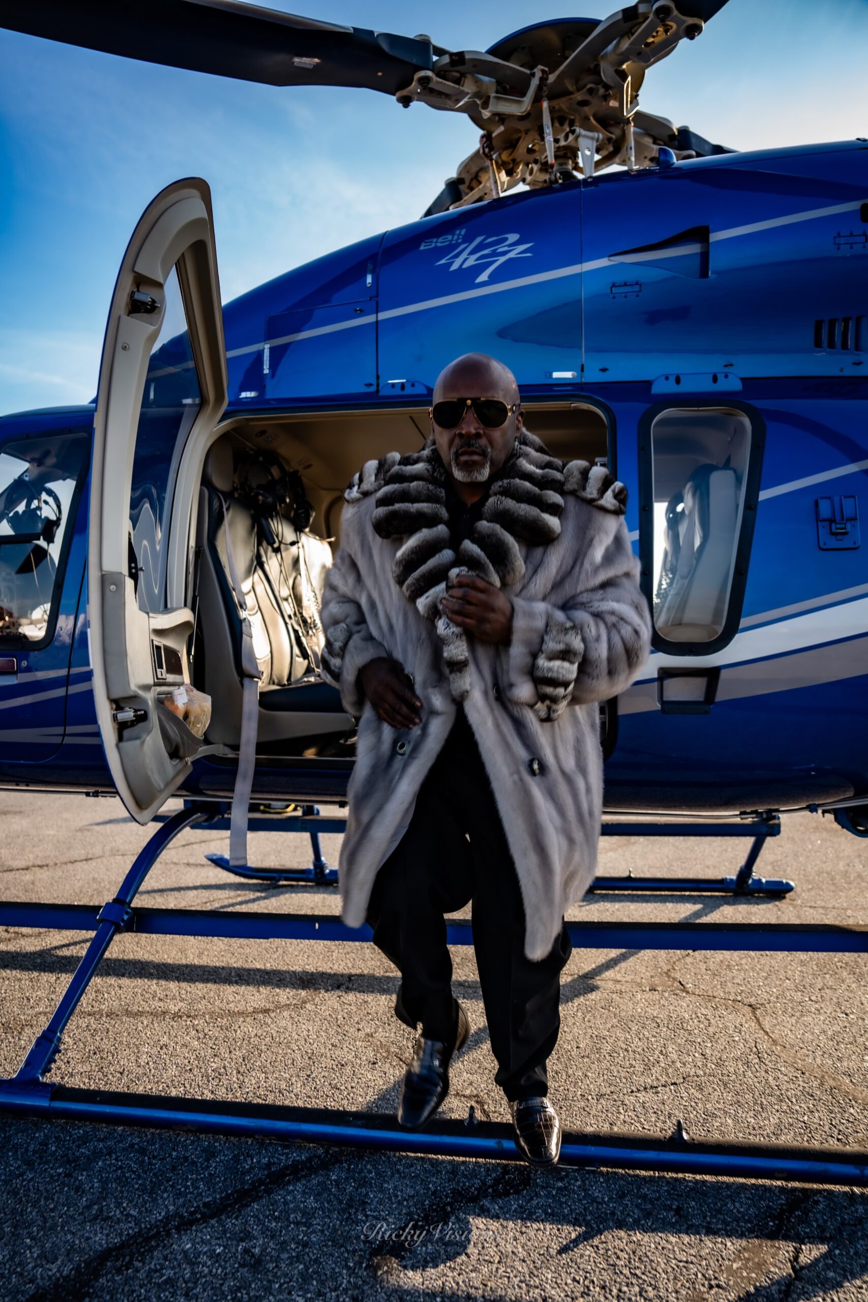 A Man Wearing a Long Fur Coat and Sunglasses Steps Out of a Blue Helicopter